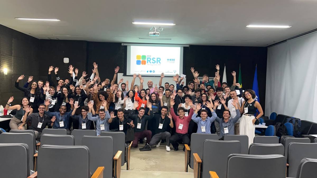 Participantes de braços erguidos em auditório, com a tela da RSR Rio ao fundo